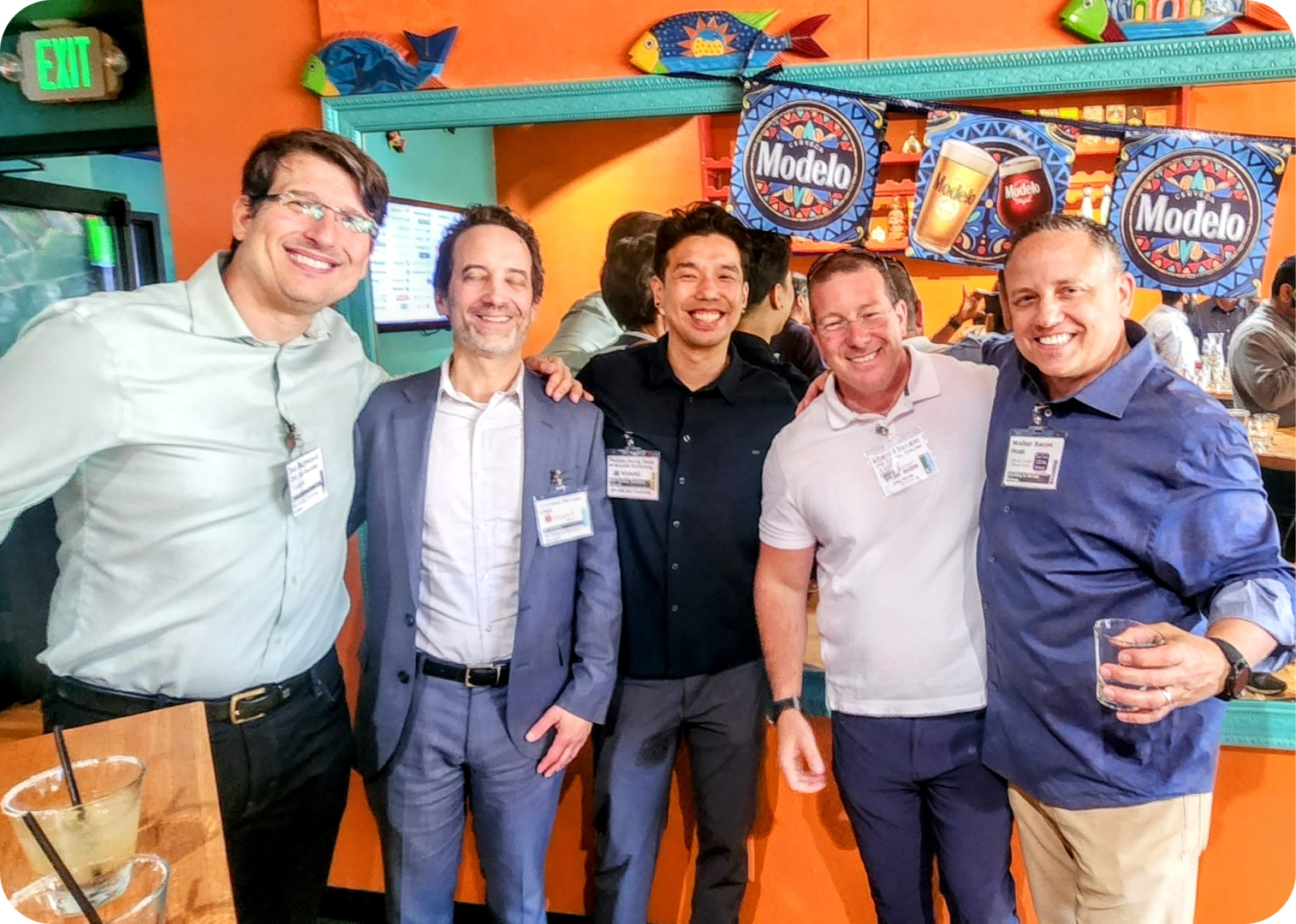 Four men smiling in front of Modelo beer signs.