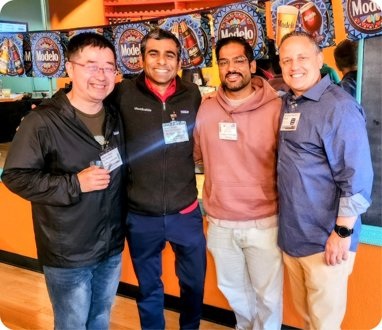 Four men smiling in front of Modelo beer signs.